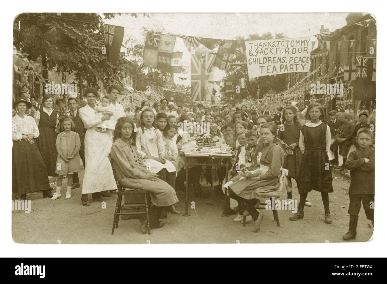 Cartolina originale del WW1 di un gruppo di bambini seduti ai tavoli all'esterno della strada per un Partito per la Pace per celebrare la fine della guerra mondiale del 1st - il banner recita ' Thanks to Tommy & Jack for Our Childrens Peace Tea', Boston Rd., Walthamstow London, Regno Unito del 1918 Foto Stock