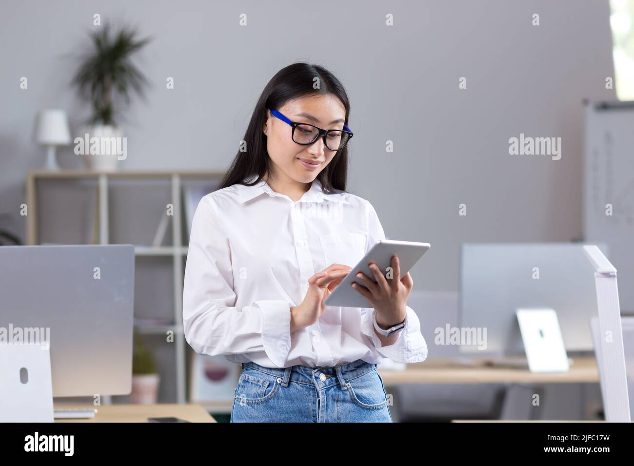 Giovane insegnante asiatico bello che lavora con il tablet, in classe con i computer a scuola, donna in camicia bianca e occhiali legge dal tablet Foto Stock