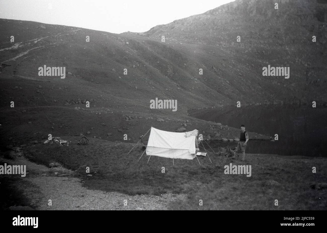 1952, storico, campeggio ad angolo tarn, Lake District, Cumbria, Inghilterra, Regno Unito, un giovane uomo in piedi fuori una tenda di tela dell'epoca con il lago di montagna alle spalle. Foto Stock
