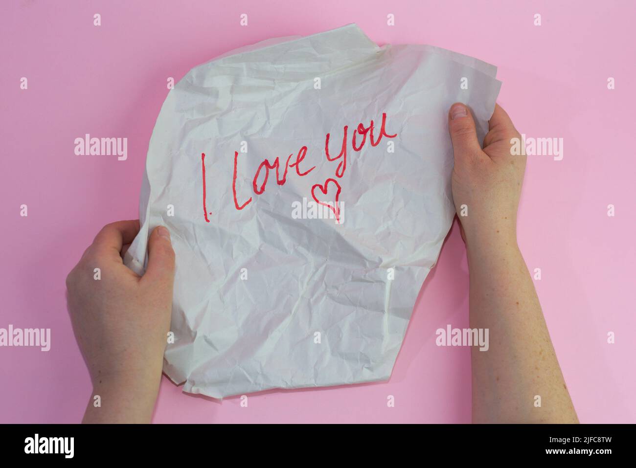Un primo piano di mani tenendo un pezzo di carta sbriciolato con un segno i Love You su uno sfondo rosa Foto Stock