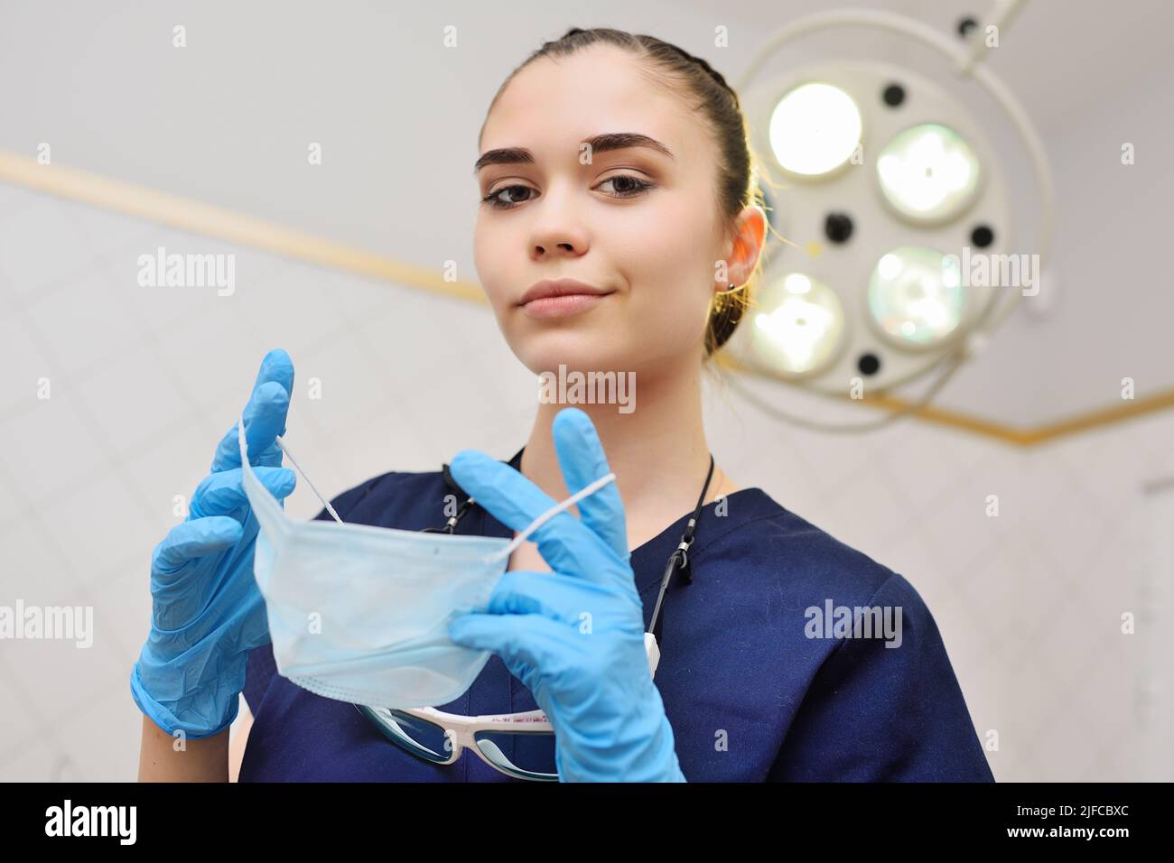 bella giovane donna in un abito chirurgico in guanti di gomma blu sullo sfondo di una lampada chirurgica mette su una maschera sterile. Foto Stock