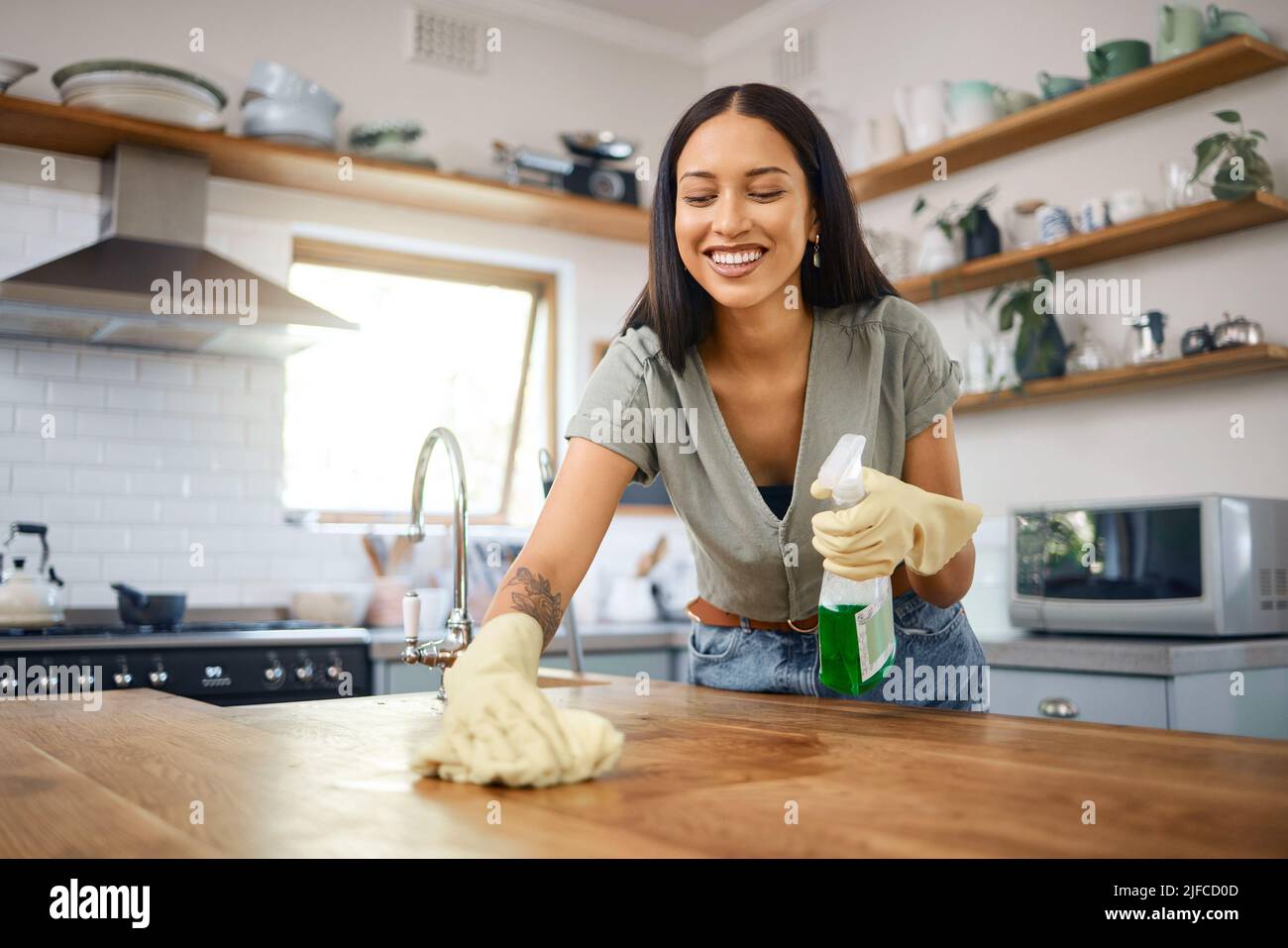 Una giovane donna da corsa mista sorridente che indossa guanti protettivi e usa una bottiglia spray con igienizzatore per pulire i suoi banchi da cucina a casa. Ispanico Foto Stock