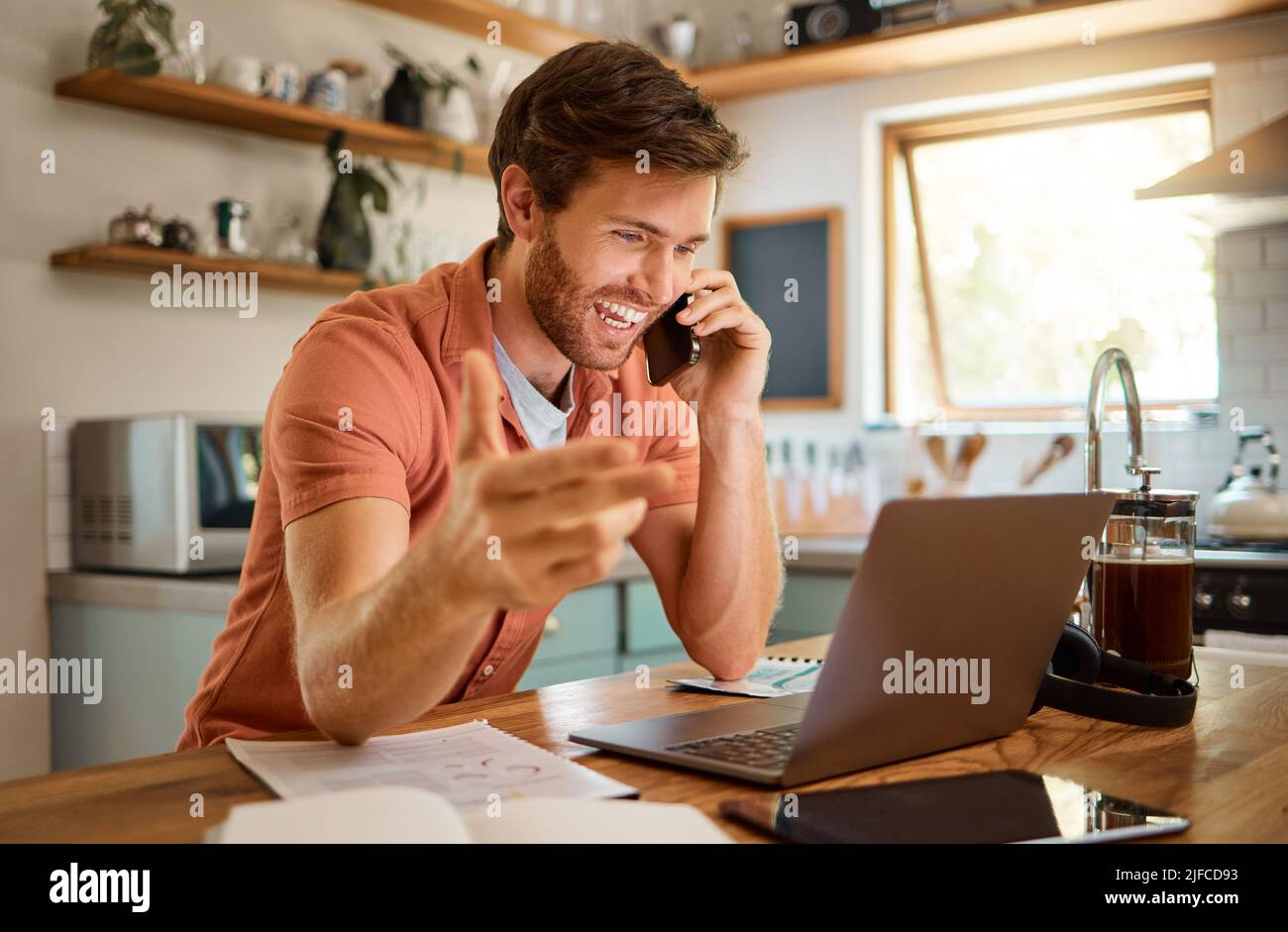 Contenuto giovane uomo d'affari caucasico su una chiamata utilizzando un telefono mentre si lavora su un portatile a casa da solo. Felice uomo d'affari maschile sorridendo e parlando Foto Stock