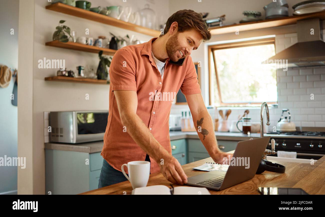 Contenuto giovane uomo d'affari caucasico su una chiamata utilizzando un cellulare mentre si lavora su un laptop e bere caffè a casa da solo. Felice uomo d'affari maschile Foto Stock