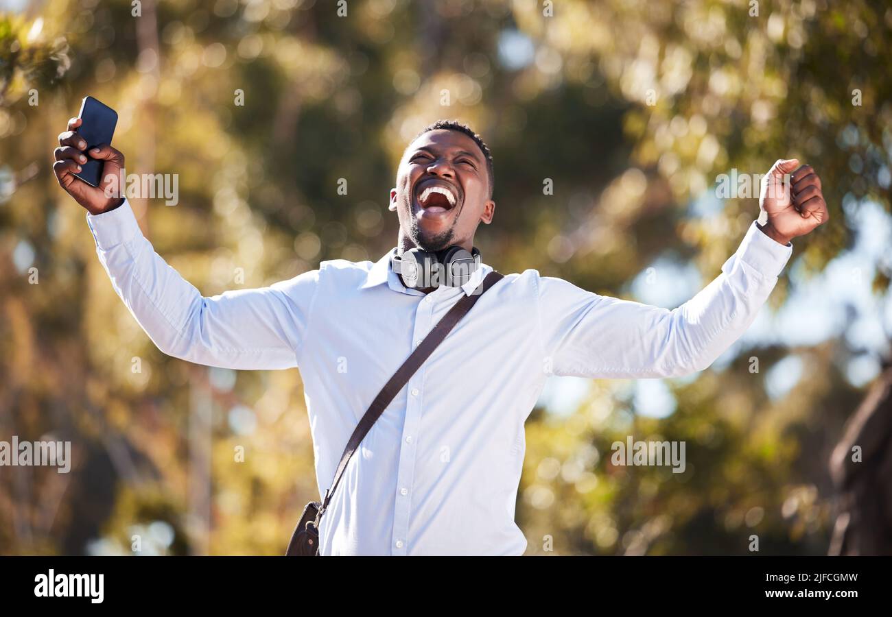 L'uomo afro-americano festeggia una vittoria mentre usa il suo cellulare e indossa le cuffie. Felice giovane maschio che esprime successo e vittoria mentre cammina Foto Stock