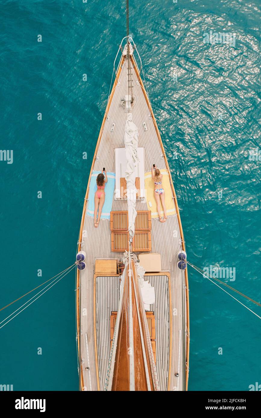 Due donne in bikini giacciono su yacht prendendo il sole utilizzando cellulari dall'alto. Amici in vacanza prendere il sole su yacht durante la crociera utilizzando cellulari a. Foto Stock