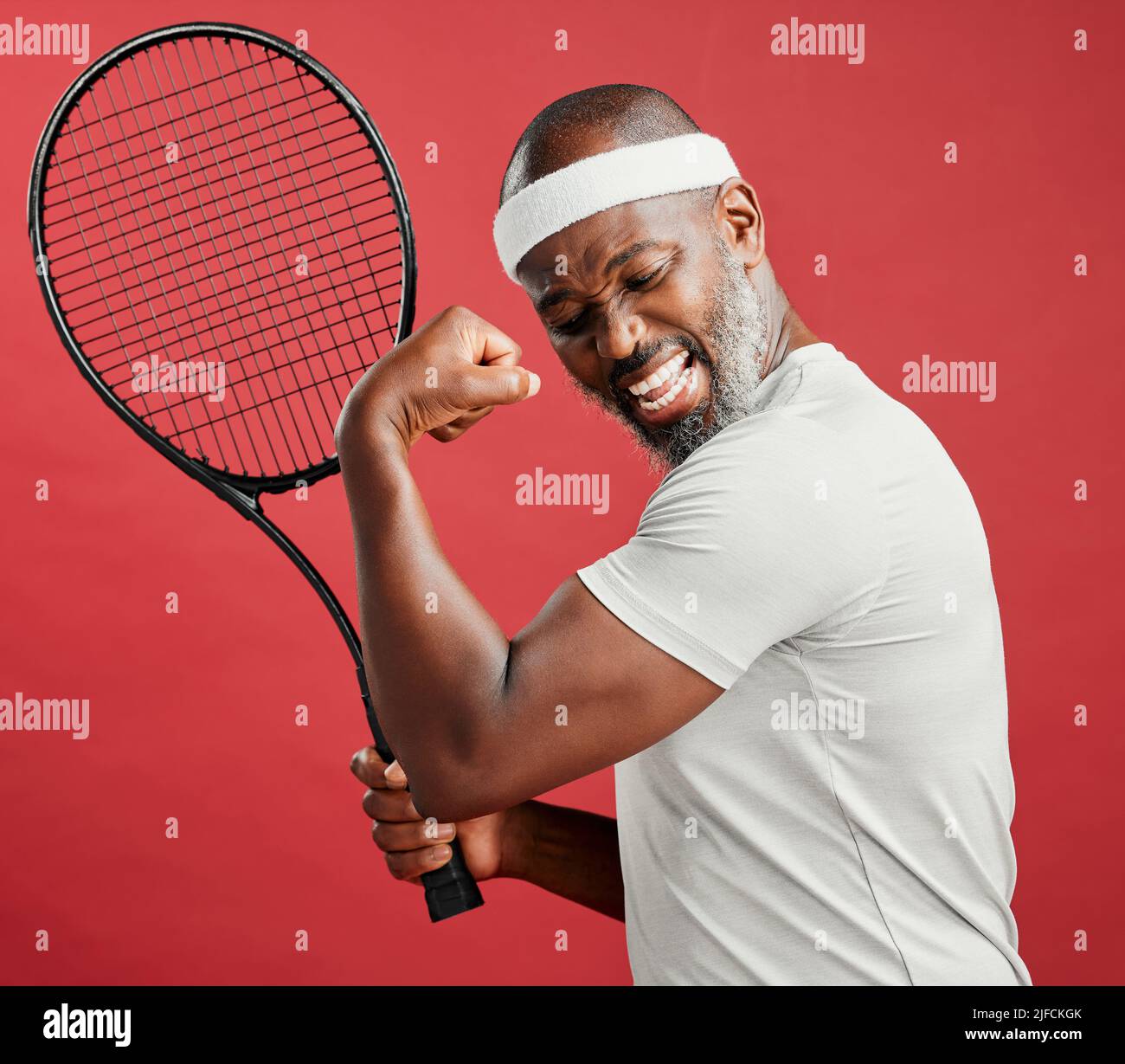 Un uomo afro-americano maturo felice in piedi su uno sfondo rosso in studio, flettendo il suo bicep mentre si posa con una racchetta da tennis. Nero sorridente Foto Stock