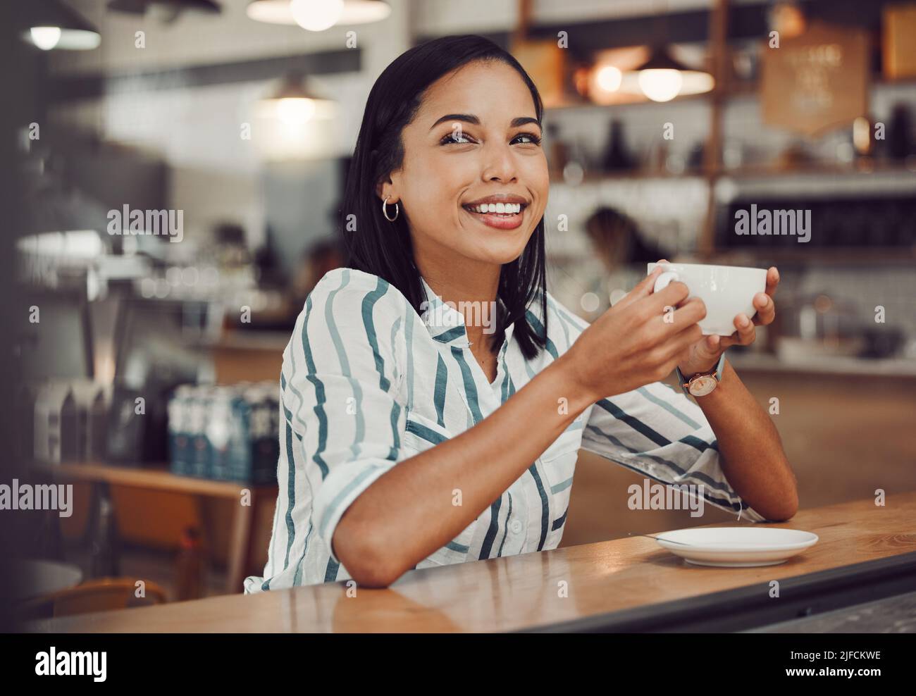 Felice giovane sorridente donna mista gara in casual vestito bere bevande calde mentre si siede da solo a un tavolo in un accogliente caffè. Bella femmina cercando Foto Stock