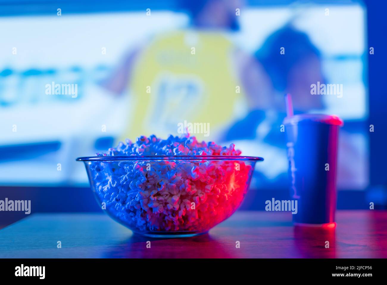 Sullo sfondo di una partita di calcio sullo schermo della TV, una grande ciotola di vetro di popcorn e una bevanda in un bicchiere. Guarda i tuoi programmi sportivi preferiti o Foto Stock