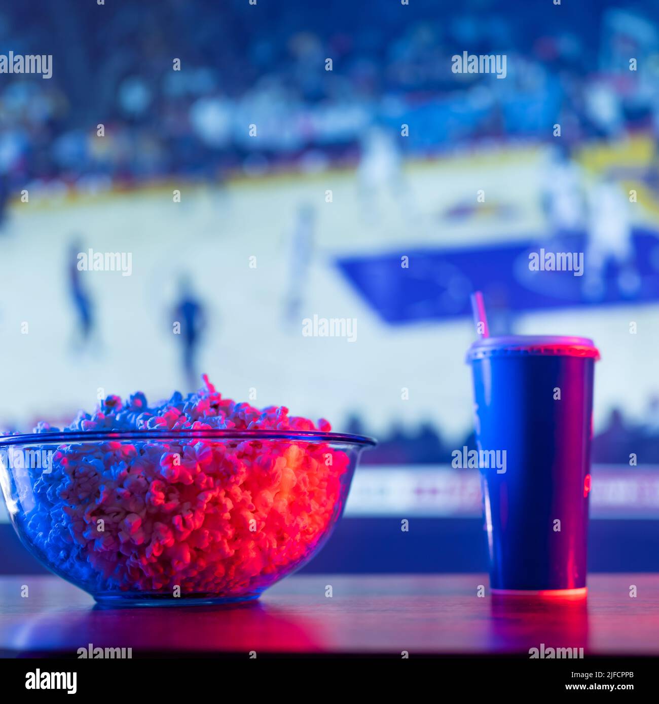 Fast food - popcorn in una ciotola di vetro, una bevanda in un bicchiere di plastica sullo sfondo di uno schermo TV con una partita sportiva. Calcio, basket, cucina americana Foto Stock