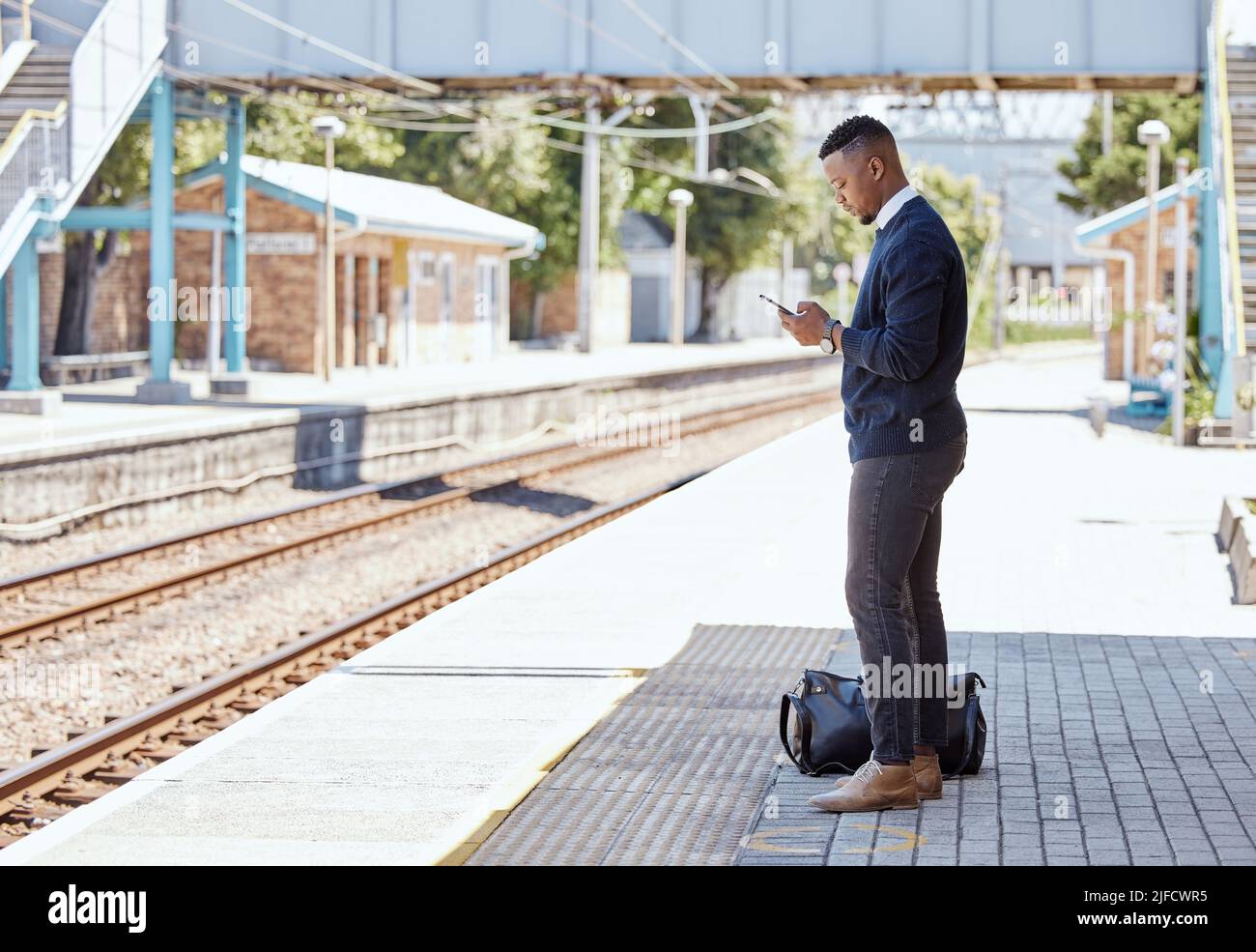 Uomo d'affari nero che viaggia da solo. Un giovane uomo d'affari afroamericano in attesa di un treno in una stazione ferroviaria e utilizzando il suo cellulare wireless Foto Stock
