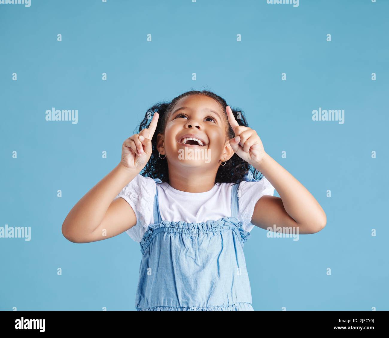 Eccitato bambino piccola ragazza che osserva stupito e sorpreso mentre punta le sue dita in su allo spazio della copia contro sfondo blu dello studio. Pubblicità bambini Foto Stock