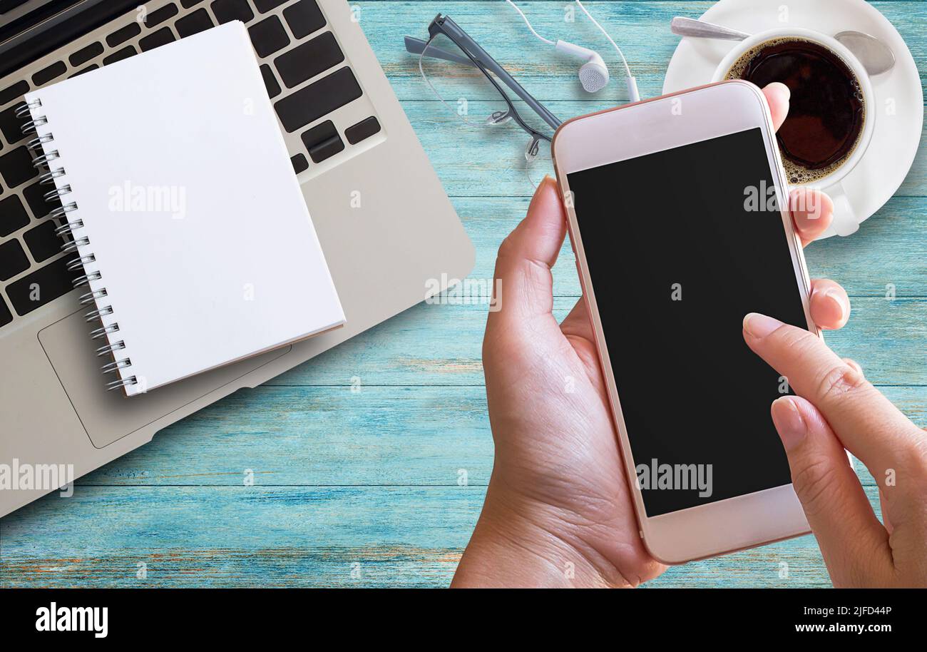 Smartphone in mano femminile con scrivania da ufficio con computer portatile, caffè e bicchieri su sfondo tavolo di legno. Luogo di lavoro. Vista dall'alto con spazio per la copia. Apprendimento Foto Stock