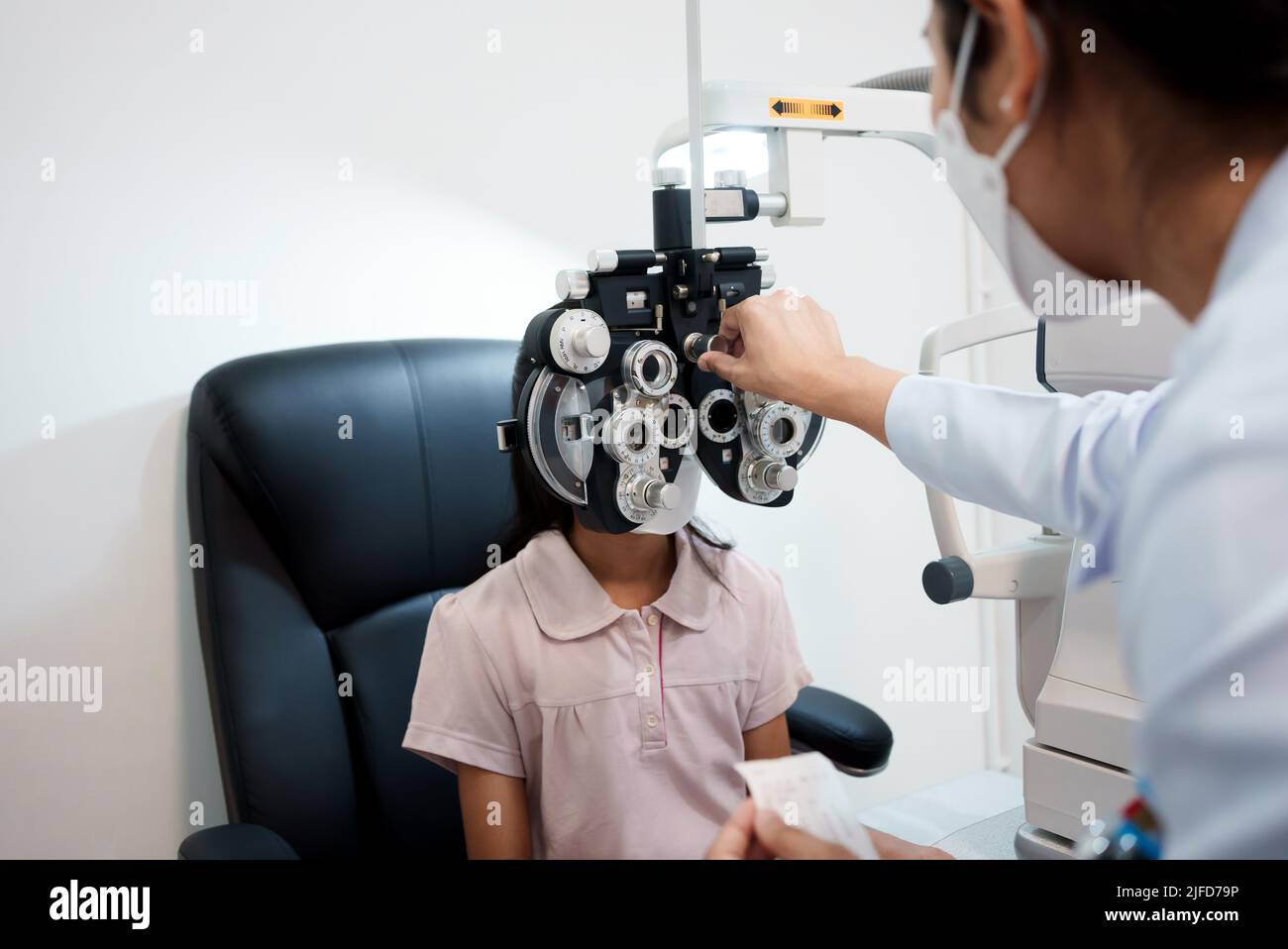 Oftalmologo che esamina gli occhi di una ragazza asiatica in una clinica. Indossano maschere protettive. Foto Stock