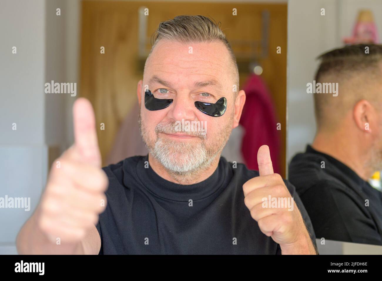 Uomo anziano che usa i cerotti idratanti dell'occhio del gel che dà i pollici doppi in su di approvazione in un concetto di cura della pelle Foto Stock