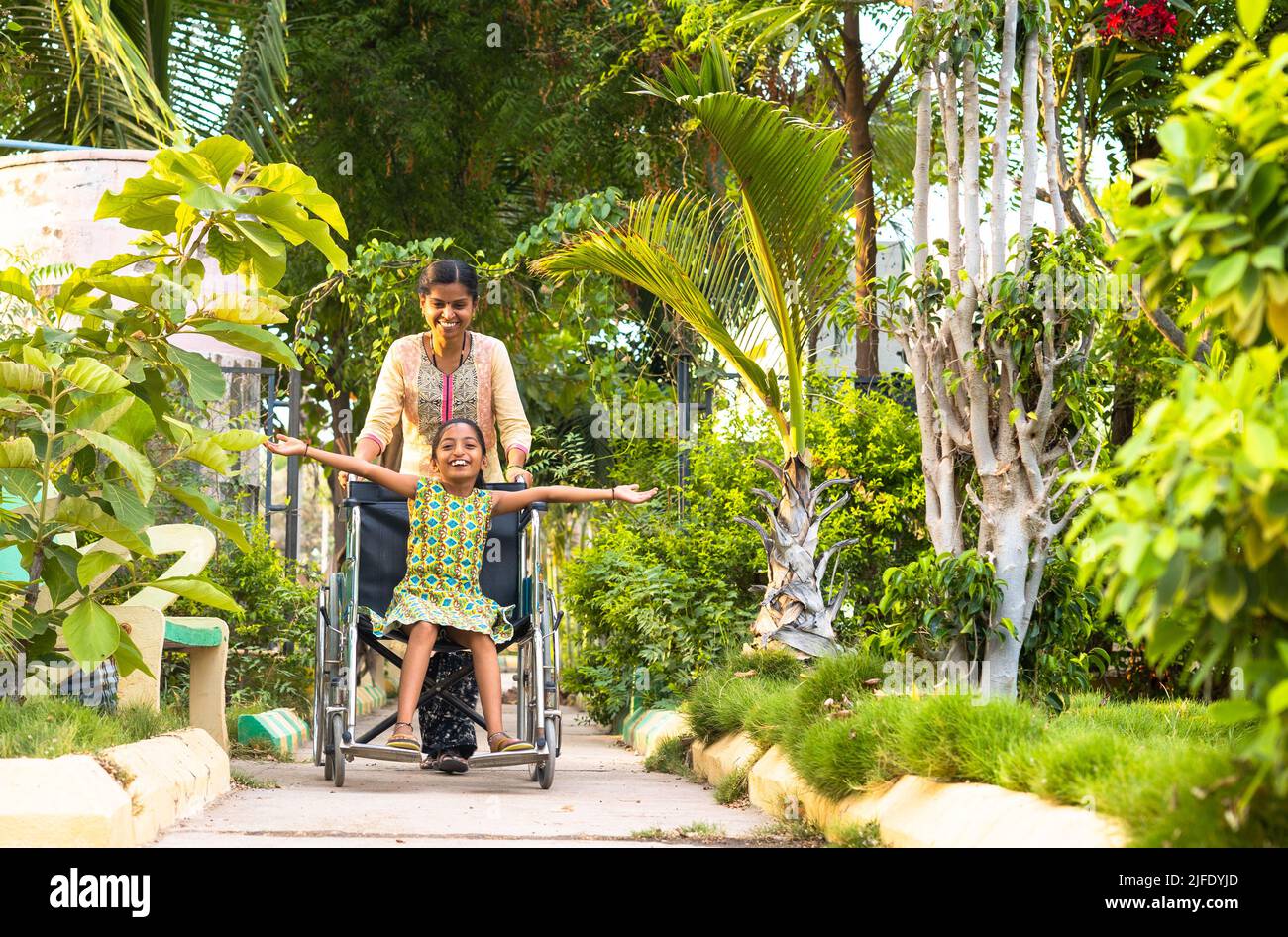 Madre che prende il bambino con disabilità a piedi mentre usa la sedia a rotelle al parco - concetto di maternità, cura e relax. Foto Stock