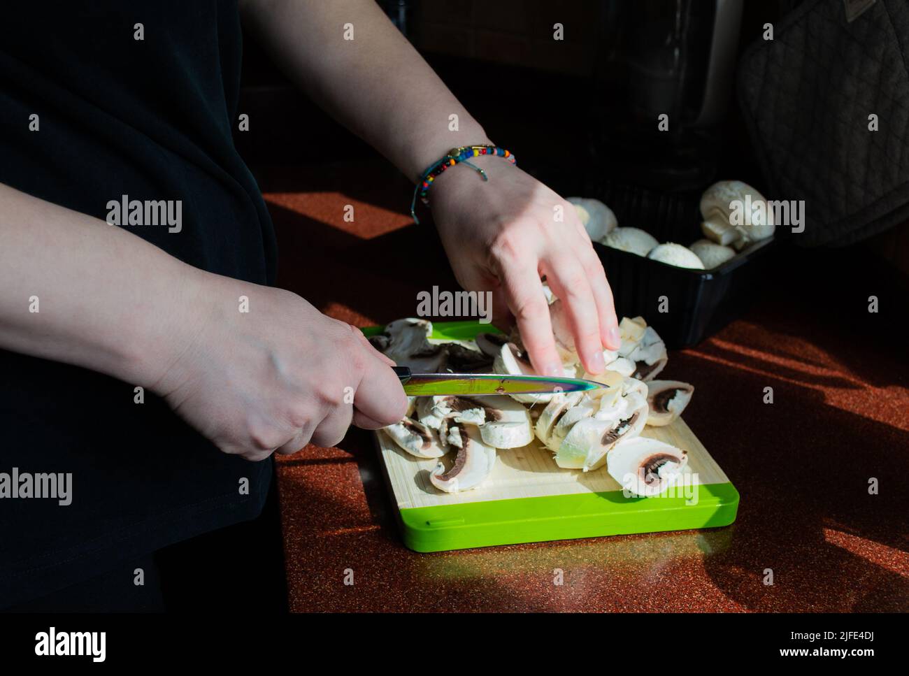 Primo piano delle mani con funghi freschi a fette su un tagliere. Cucina vegetariana fatta in casa. Luce solare dalla finestra, ombre sul tavolo. Foto Stock