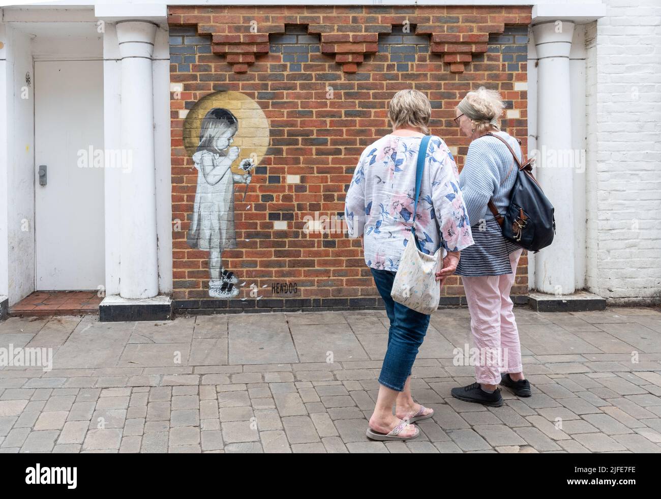 Due Signore che ammirano il murale dell'arte di strada di Hendog, un artista anonimo, raffigurante una giovane ragazza che raccoglie i petali di un fiore, a Winchester, Hampshire, Regno Unito. Foto Stock