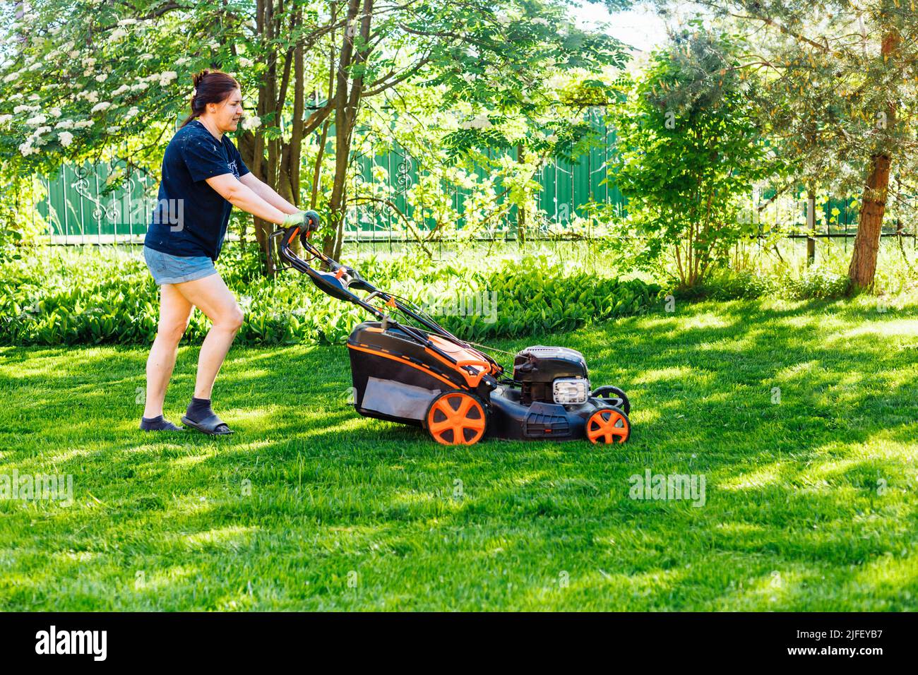 Giardiniere femmina dai capelli castani in abiti casual con rasaerba a benzina o tagliaerba in giardino. Attività nel fine settimana Foto Stock