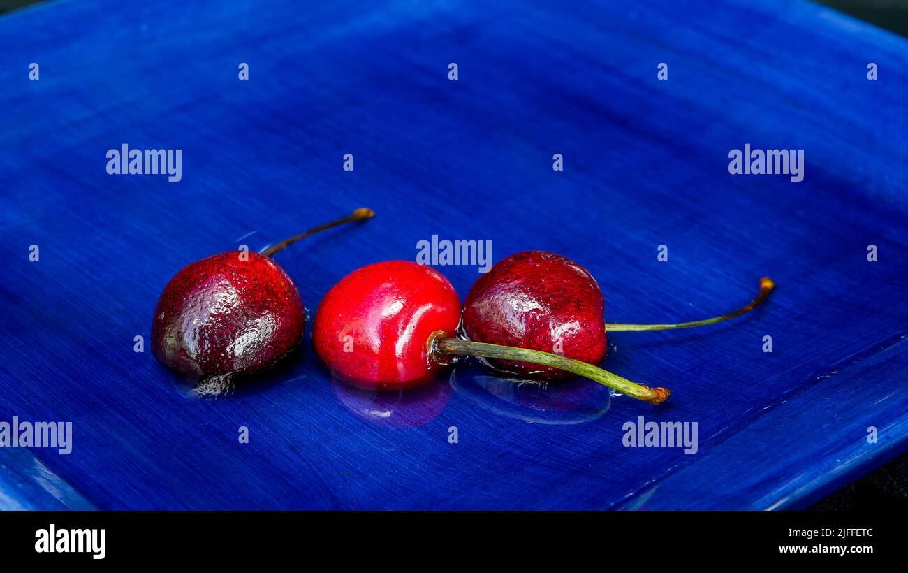 Tre ciliegie rosse su piastra blu con acqua e spazio per la copia Foto Stock