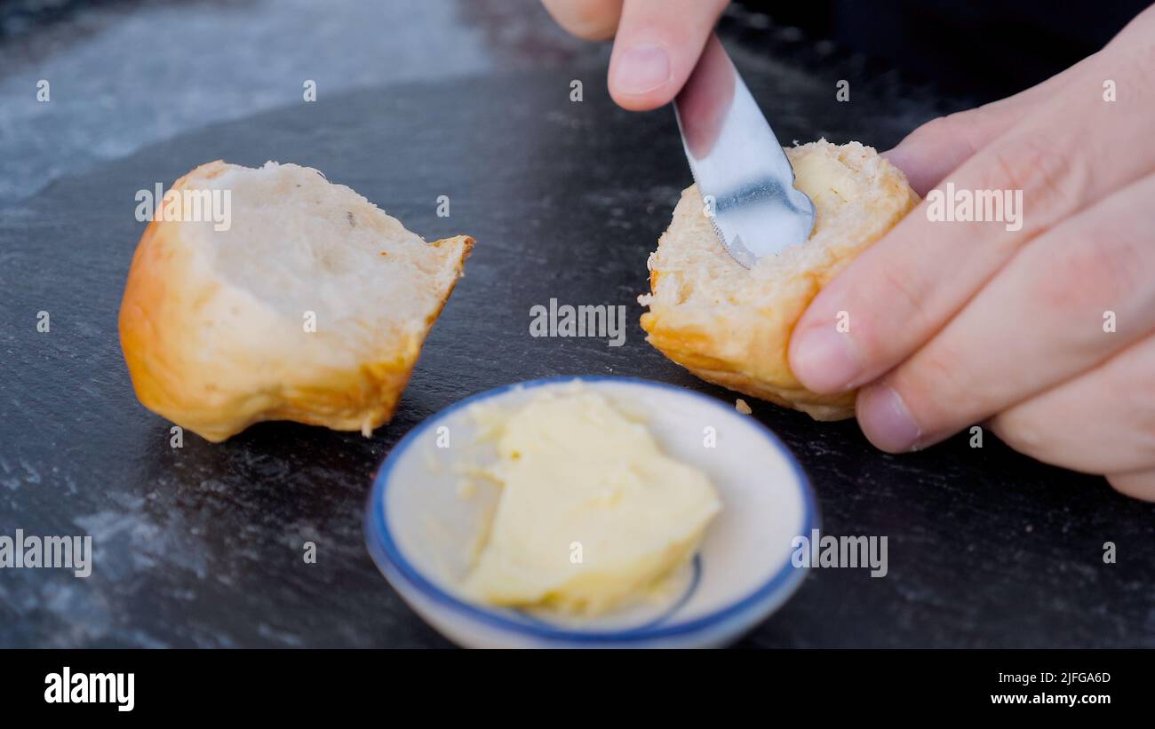 Maschio mano spalmando il burro su pane bianco con coltello in acciaio su tavola in pietra nera sul tavolo da cucina. Uomo chef che tiene fetta di fresco rustico bun Foto Stock