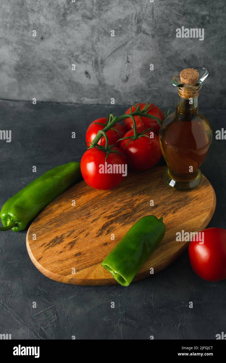 Cibo ancora vivo con olio d'oliva, peperoni verdi e pomodori maturi in scena scura. Vista ad angolo elevato delle verdure fresche con tavoletta di legno e spazio per la copia. Foto Stock