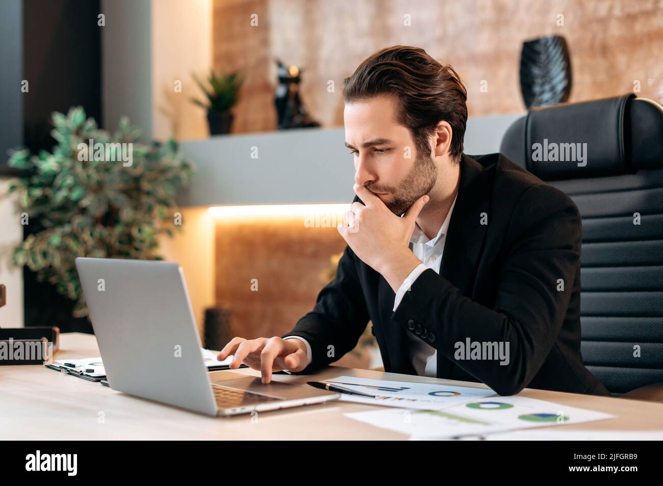Un uomo d'affari caucasico, un manager, un leader aziendale, si siede sul posto di lavoro in ufficio, guarda con attenzione allo schermo del laptop, lavora su un piano aziendale per un nuovo progetto Foto Stock