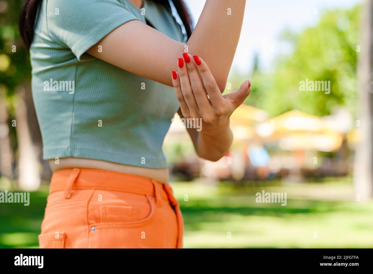 Giovane donna brunetta con tee turchese e corteccia arancione sul parco cittadino, all'aperto affetti da reumatismi cronici articolari. Dolore al gomito e trattamento c Foto Stock