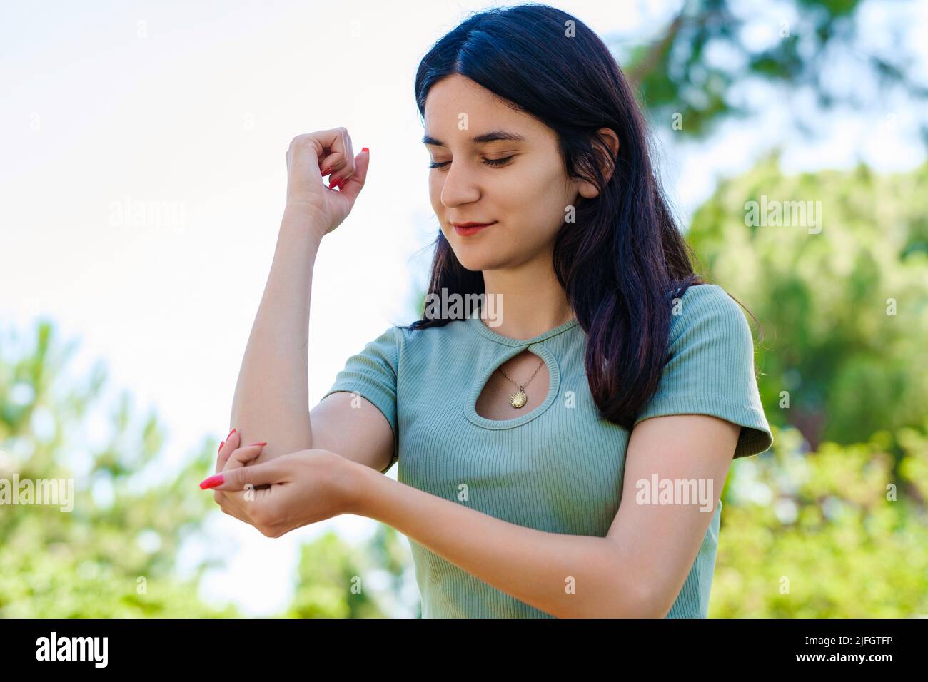 Donna bruna carina con t-shirt turchese in piedi sul parco della città, all'aperto affetti da cronico reumatismo articolare. Dolore al gomito e trattamento concep Foto Stock