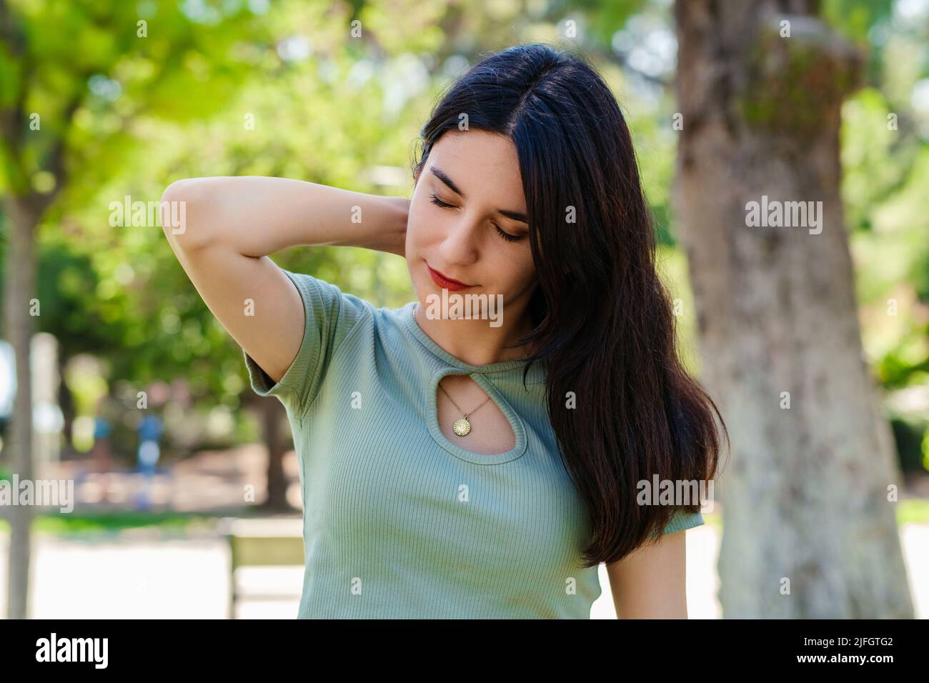 Bella donna bruna con t-shirt turchese in piedi sul parco della città, all'aperto sensazione di ferita alle spalle articolare dolore alla schiena, concetto di fibromialgia, c Foto Stock