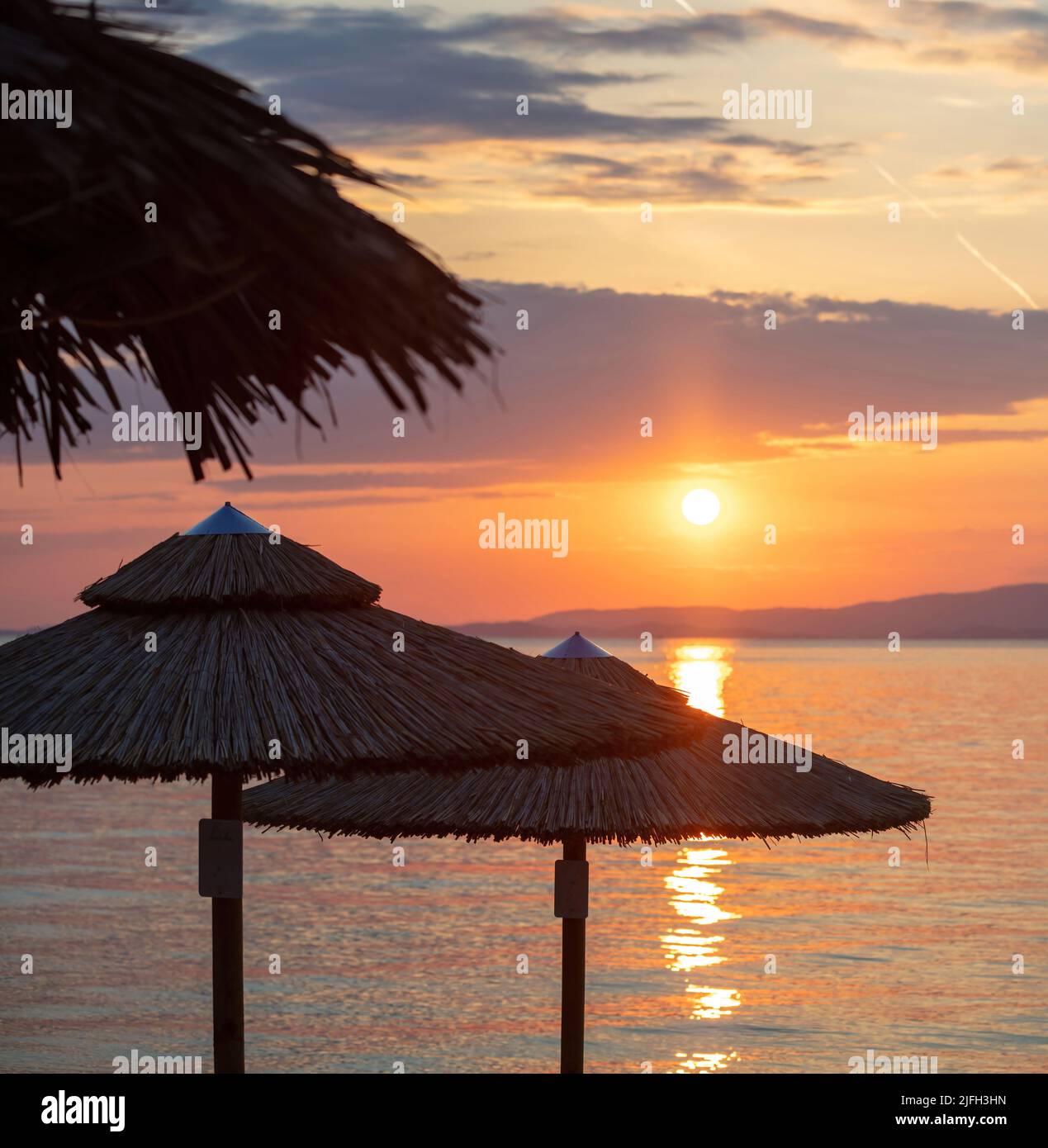 Tramonto sulla spiaggia sul Mar Egeo, Grecia. Silhouette di ombrelloni di paglia, riflessi dorati sull'acqua dell'oceano increspata, cielo di colore arancione e nuvole Foto Stock