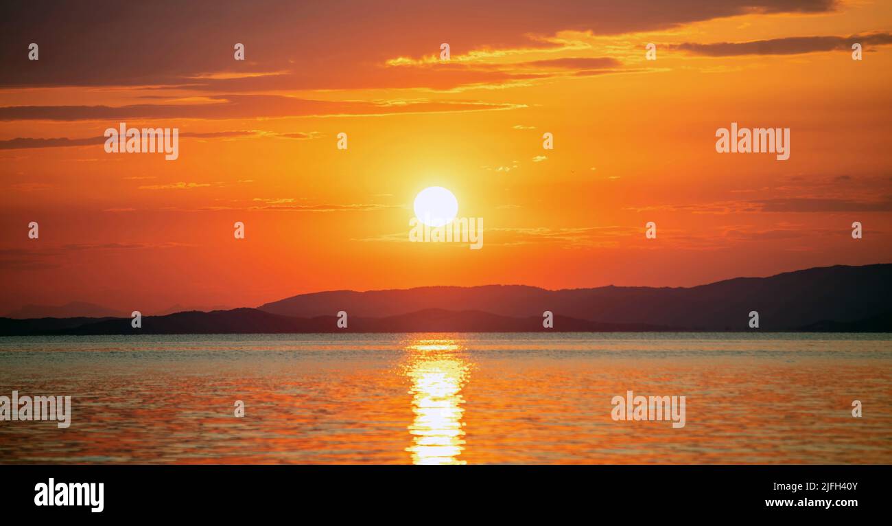 Tramonto sul Mar Egeo. Grecia. Sole che cade dietro terra scura, riflessi dorati su ondulato acqua dell'oceano. Cielo e nuvole di colore arancione Foto Stock