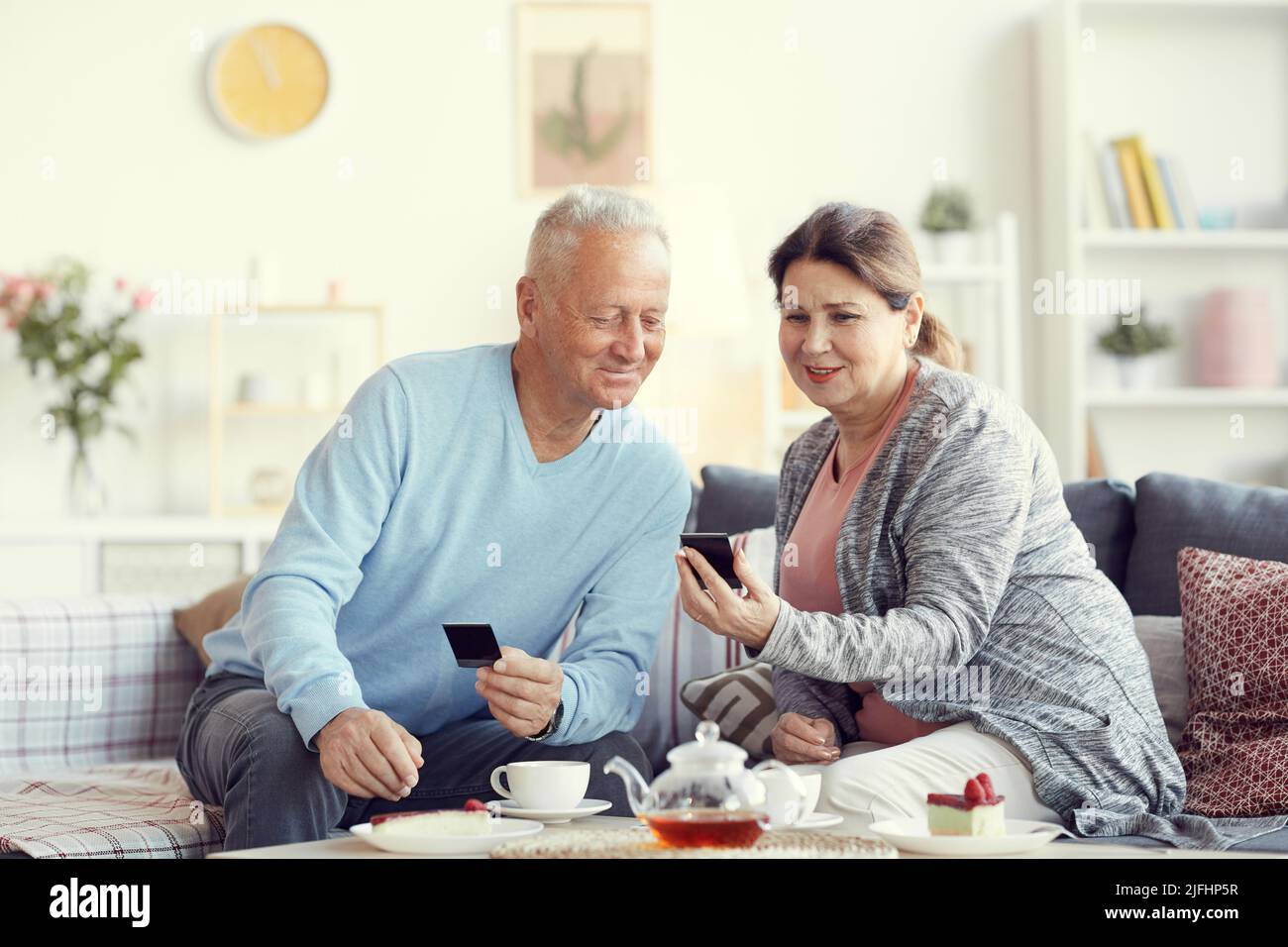 Donna anziana positiva in cardigan seduta sul divano in soggiorno e guardando vecchie foto con marito sopra la tazza di tè Foto Stock