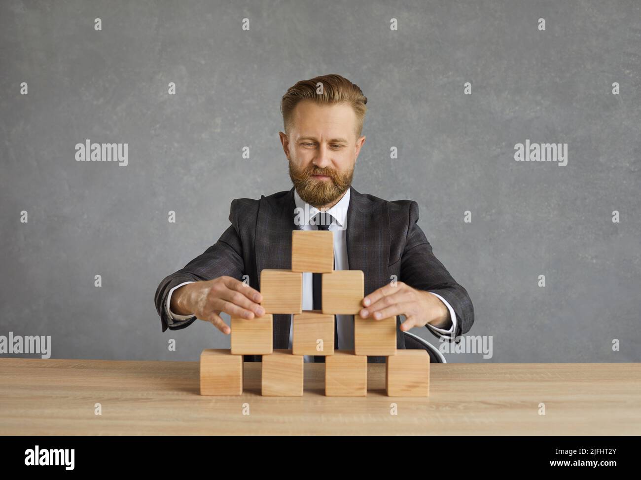 Il capo d'affari siede alla scrivania dell'ufficio e costruisce la piramide in blocchi di legno Foto Stock