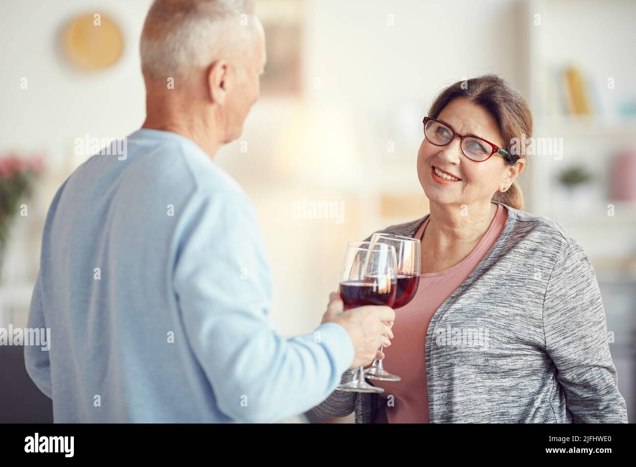 Coppia senior positiva in abbigliamento casual in piedi in soggiorno e tostatura con i wineglasses durante la celebrazione dell'anniversario Foto Stock