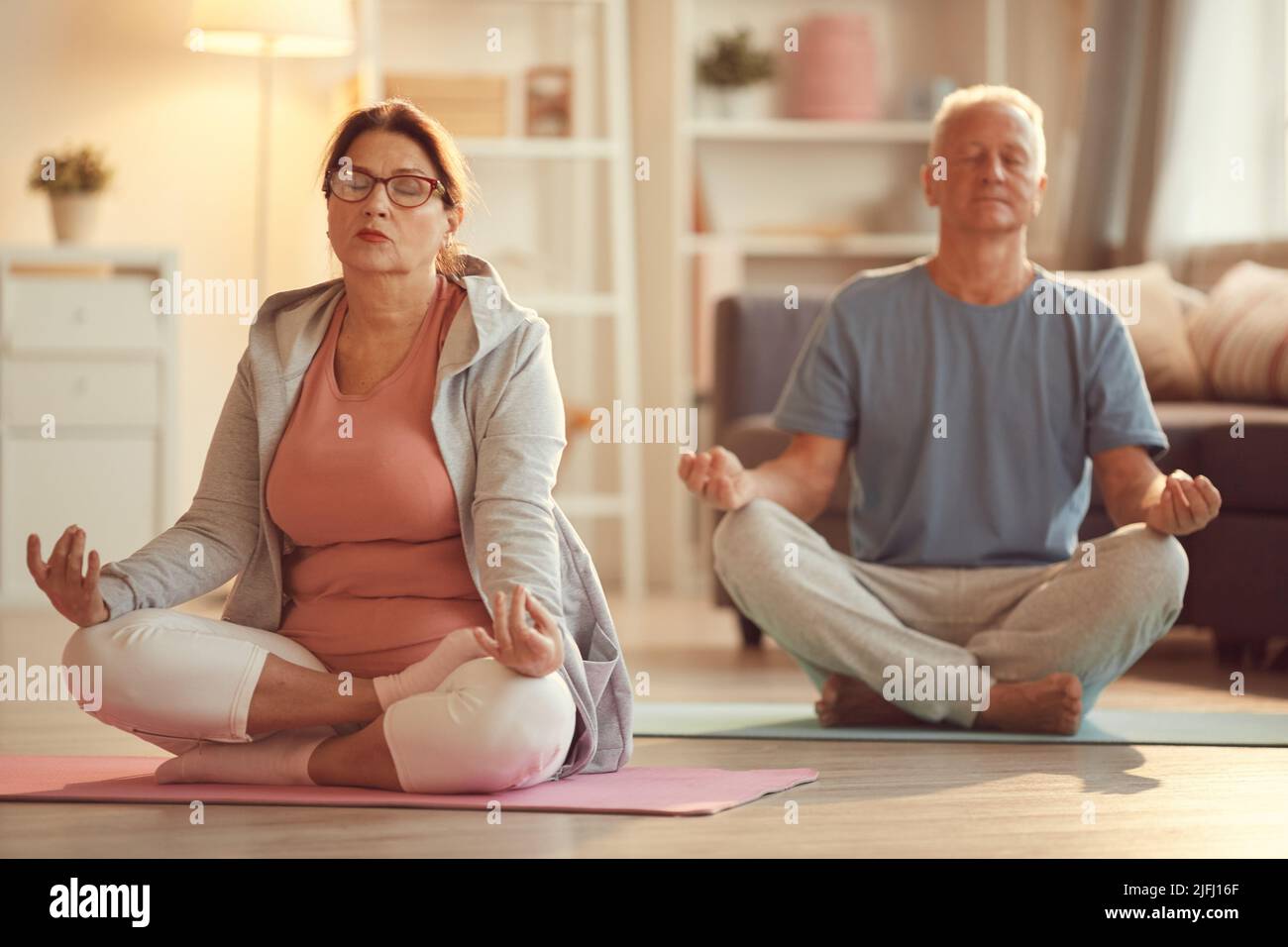Coppia senior serena e rilassata con occhi chiusi seduti con gambe incrociate sul pavimento in soggiorno e meditazione pratica Foto Stock