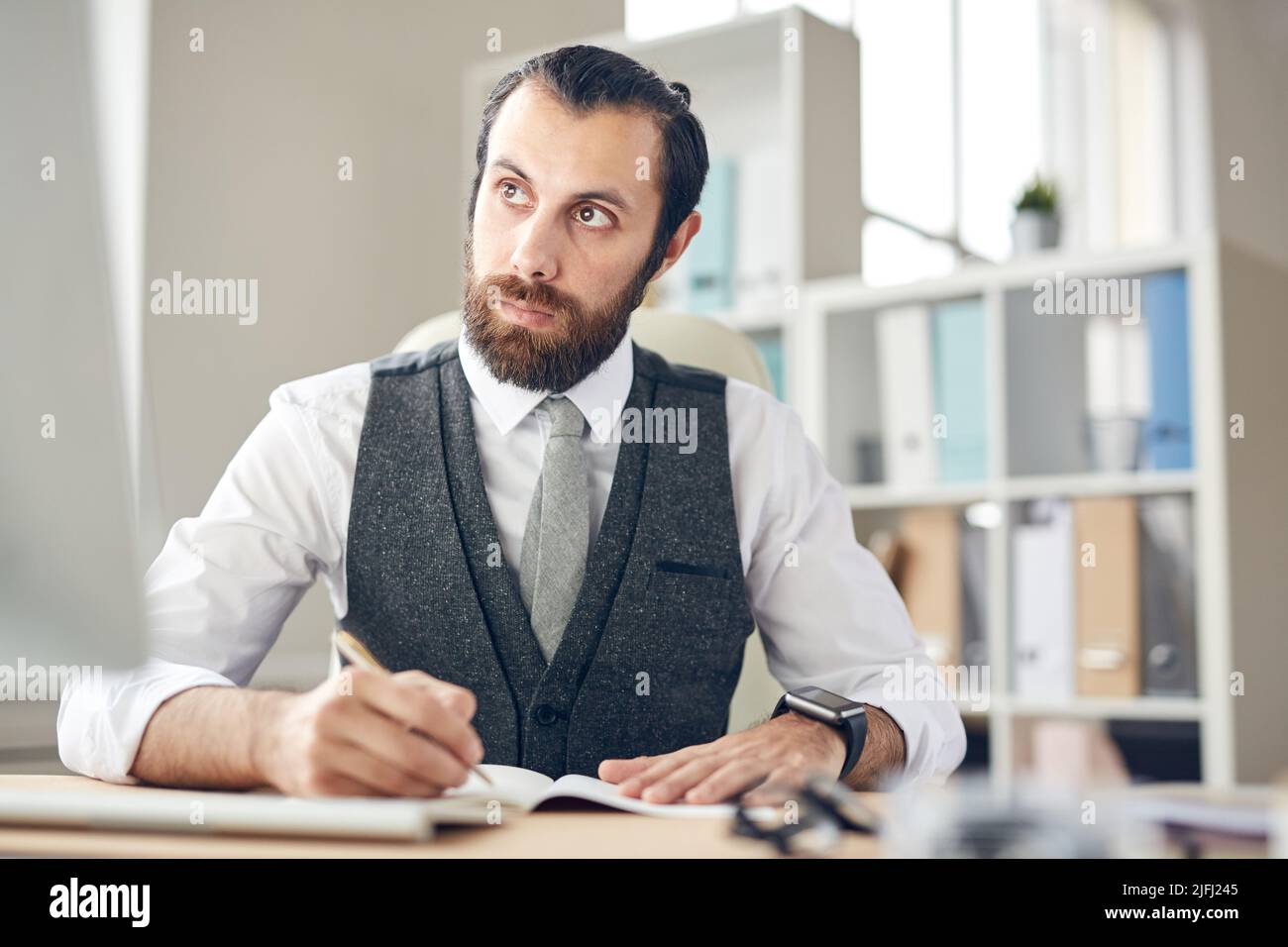 Serio giovane avvocato serio e premuroso in gilet grigio e camicia bianca seduta al tavolo e prendere appunti in pianificatore mentre si guarda computer monito Foto Stock