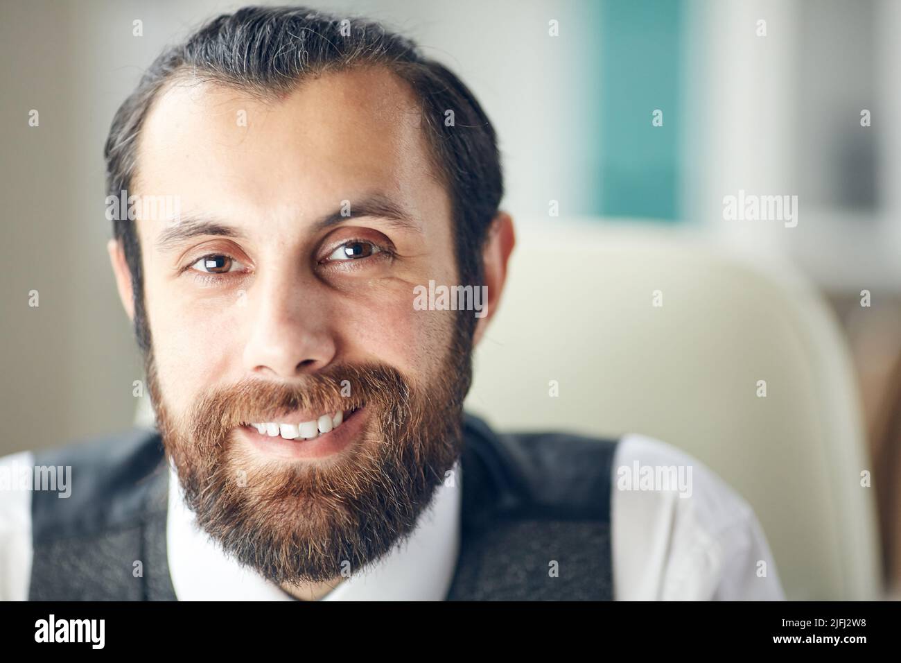Ritratto di bello e positivo giovane uomo alla moda con barba marrone e baffi sorridenti allegri in ufficio Foto Stock