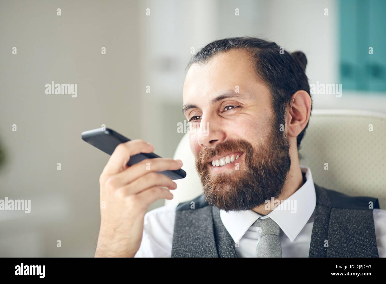 Un uomo d'affari bello e positivo seduto sulla sedia da ufficio e utilizzando lo smartphone mentre registra un divertente messaggio per il collega Foto Stock