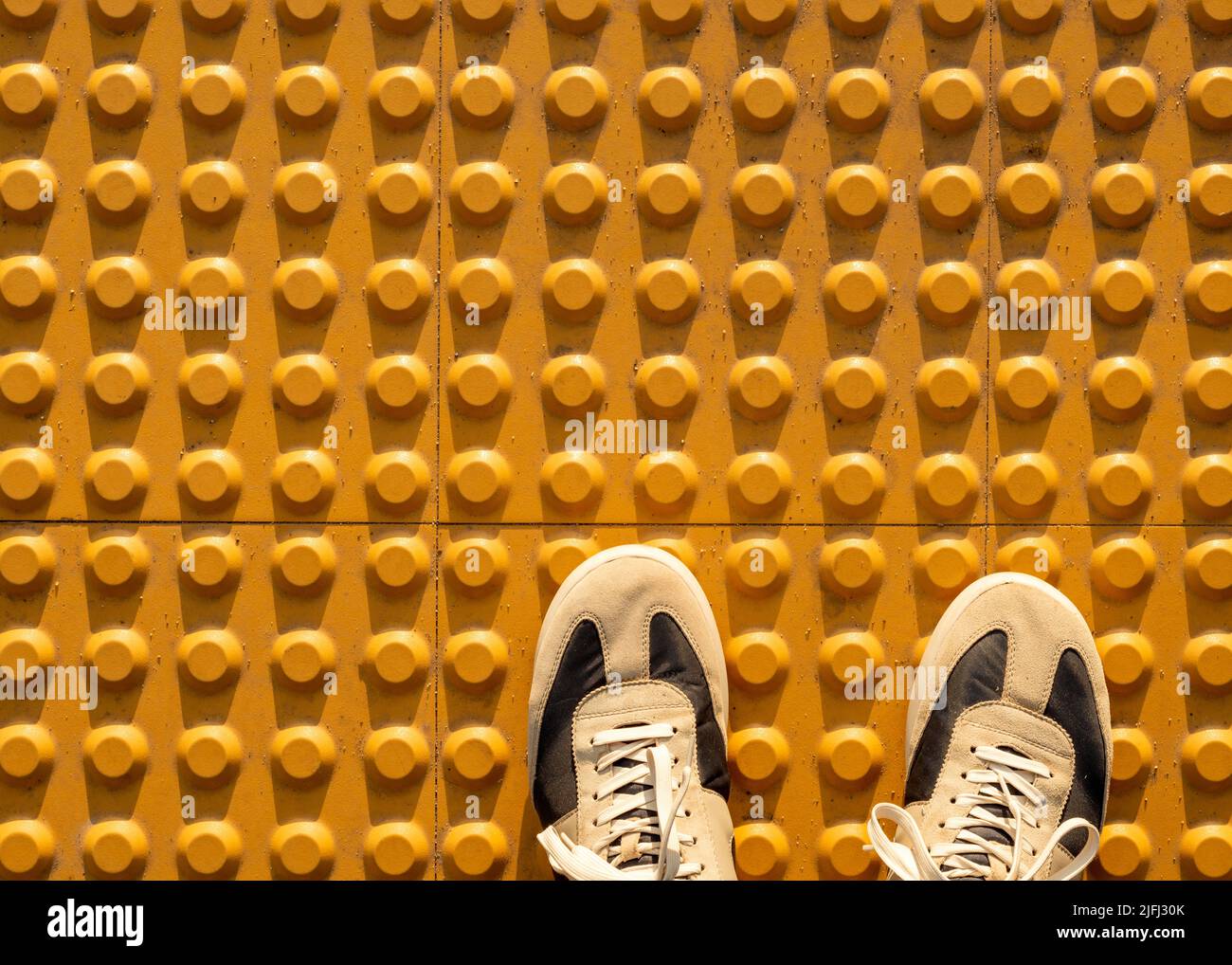 Vista dall'alto delle scarpe da ginnastica su un set di cupole troncate gialle o pavimentazione tattile gialla progettata per aiutare i pedoni con problemi di vista Foto Stock