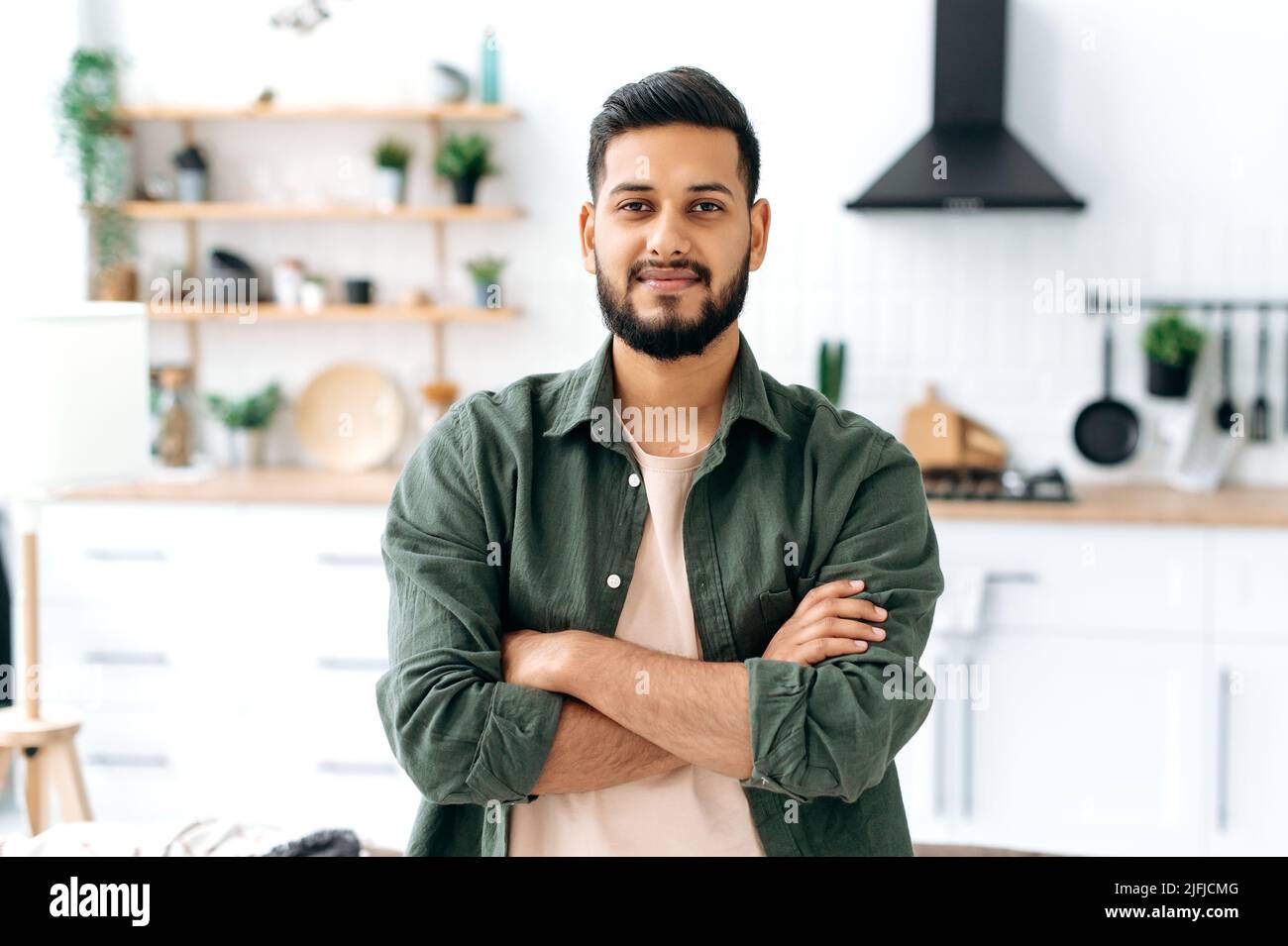 Bello fiducioso attraente indiano moderno o ragazzo arabo, in abiti casual elegante, in piedi a casa nel soggiorno sullo sfondo della cucina, con le braccia incrociate, guarda la macchina fotografica, sorridente Foto Stock
