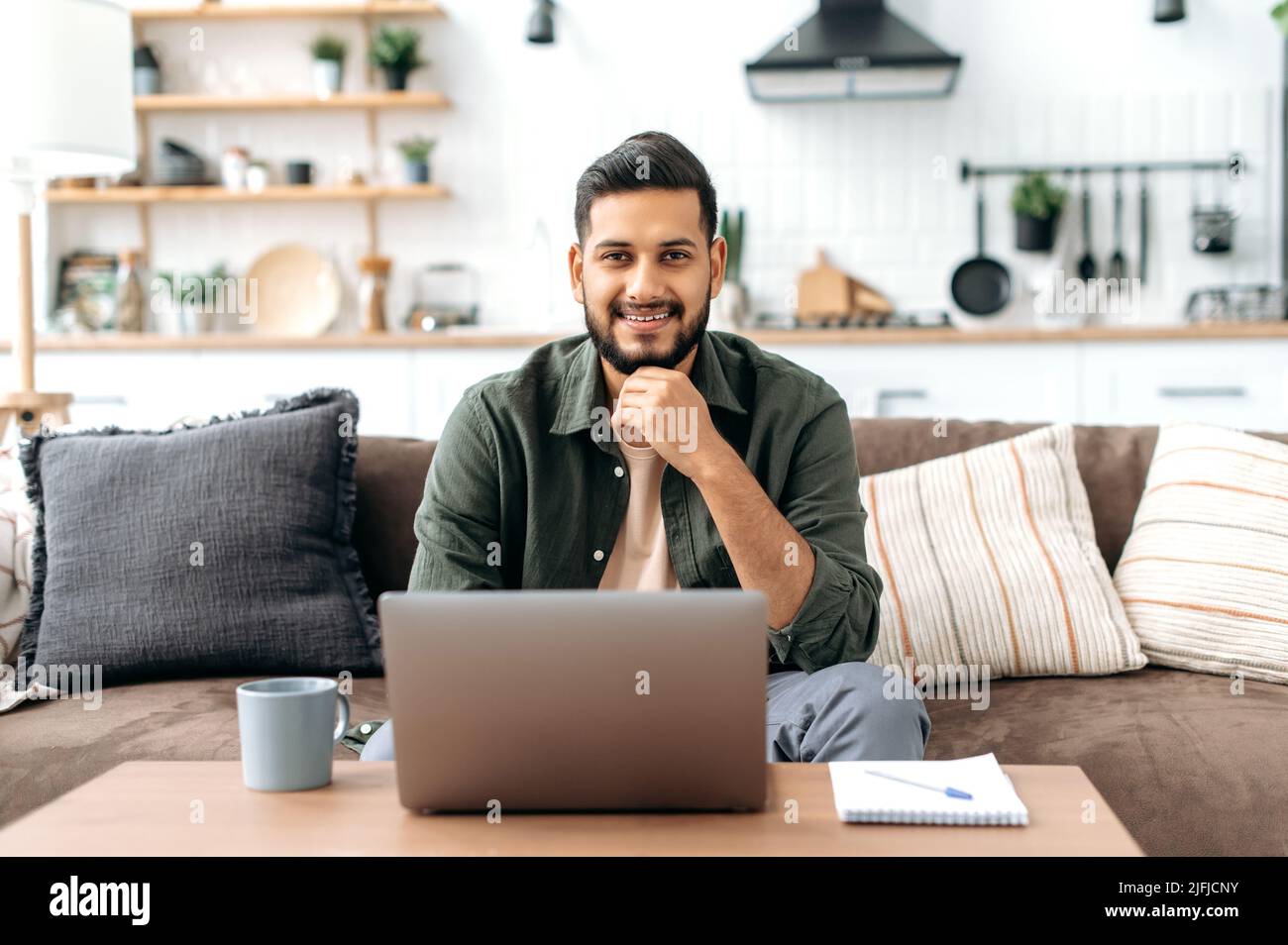 Bello sicuro attraente positivo indiano o arabo giovane uomo, in abiti casual elegante, sedersi a casa nel soggiorno sul divano, con computer portatile, guarda la macchina fotografica, sorridente amichevole Foto Stock