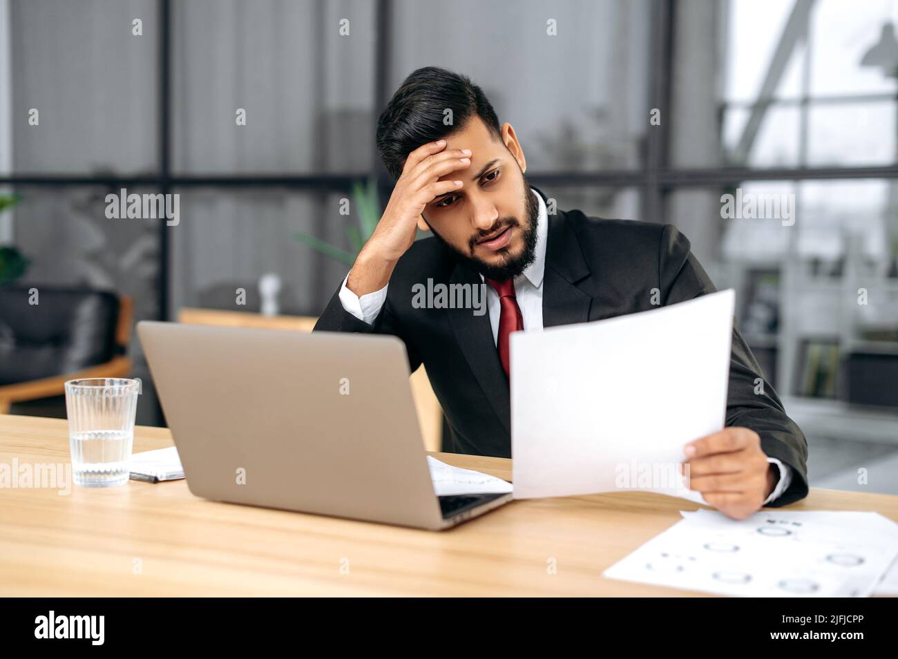Stressato uomo d'affari indiano o arabo, seduta sconvolto ad una scrivania con computer portatile in un ufficio moderno, lavora con documenti, fa un rapporto finanziario, analizza il risultato del lavoro, si sente sovraccaricato Foto Stock