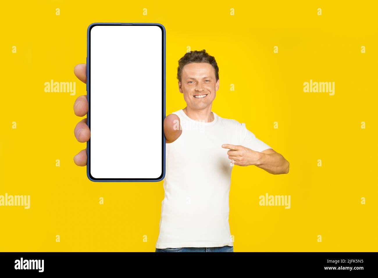 Bell'uomo biondo che punta a smartphone gigante con schermo bianco vuoto, indossando maglietta bianca e jeans display cellulare mock up isolato su sfondo giallo. Pubblicità app mobile. Foto Stock