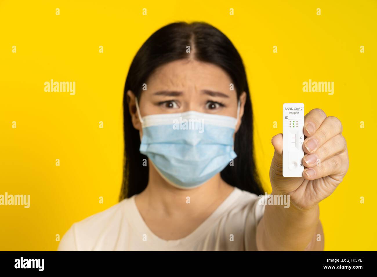 Mostra test positivo spaventato di pandemica donna asiatica matura indossando maschera medica coronavirus o prevenzione scimmipox. Donna di mezza età in maglietta bianca e maschera medica su sfondo giallo. Foto Stock