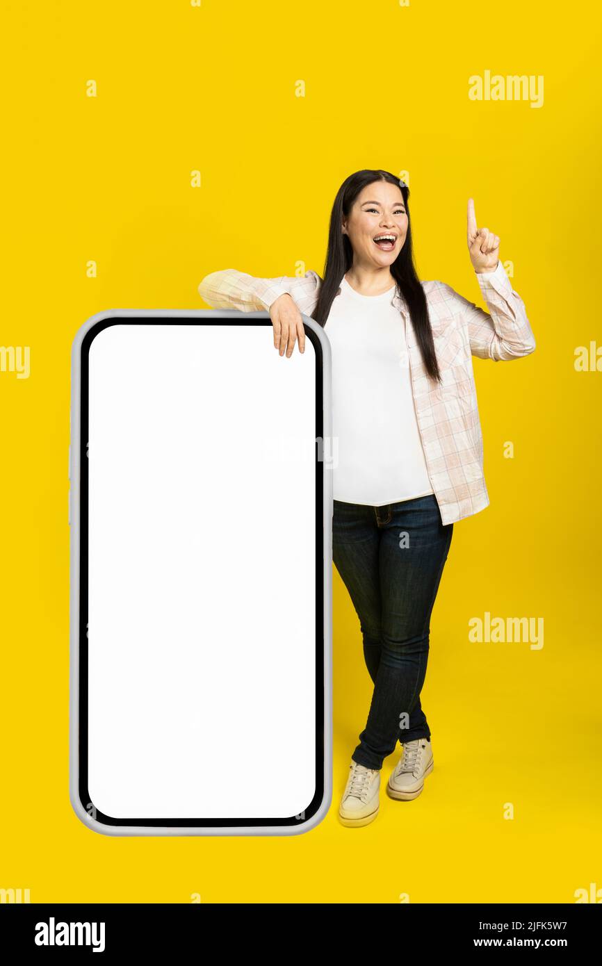 Donna asiatica di mezza età eccitata puntando il dito su appoggiata su enorme smartphone gigante con schermo bianco vuoto, indossando casual abito isolato su sfondo giallo. Spazio libero mock up. Foto Stock