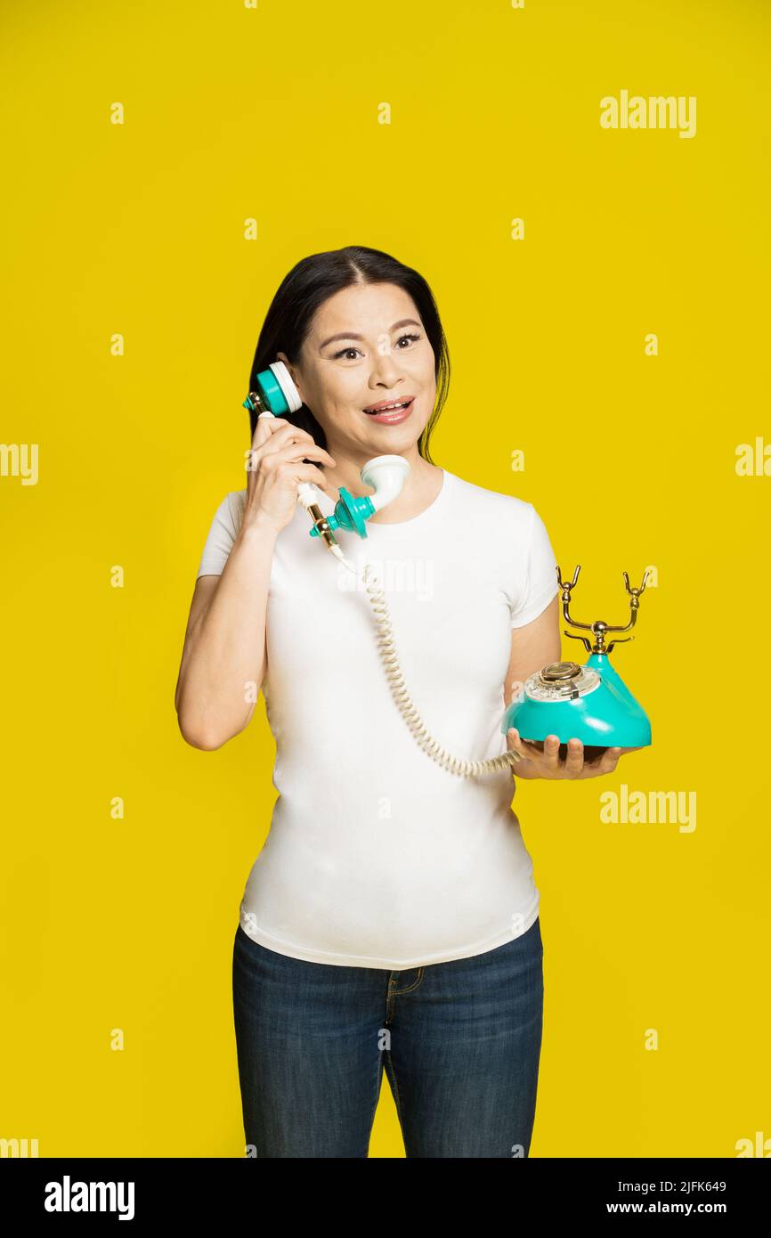 Affascinante donna asiatica divertente utilizzando vintage, telefono retrò in mani che indossano t-shirt bianca isolato su sfondo giallo. Donna asiatica eccitata con telefono retrò. Concetto di comunicazione. Foto Stock