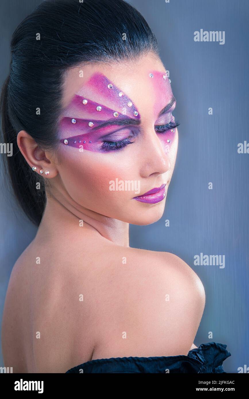 Bella donna caucasica con la pittura viola del viso e cristalli sul suo volto che guarda sopra la spalla su uno sfondo blu Foto Stock