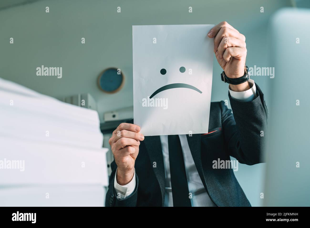 Un uomo d'affari infelice che tiene carta con emoticon accigliato in ufficio interno, fuoco selettivo Foto Stock