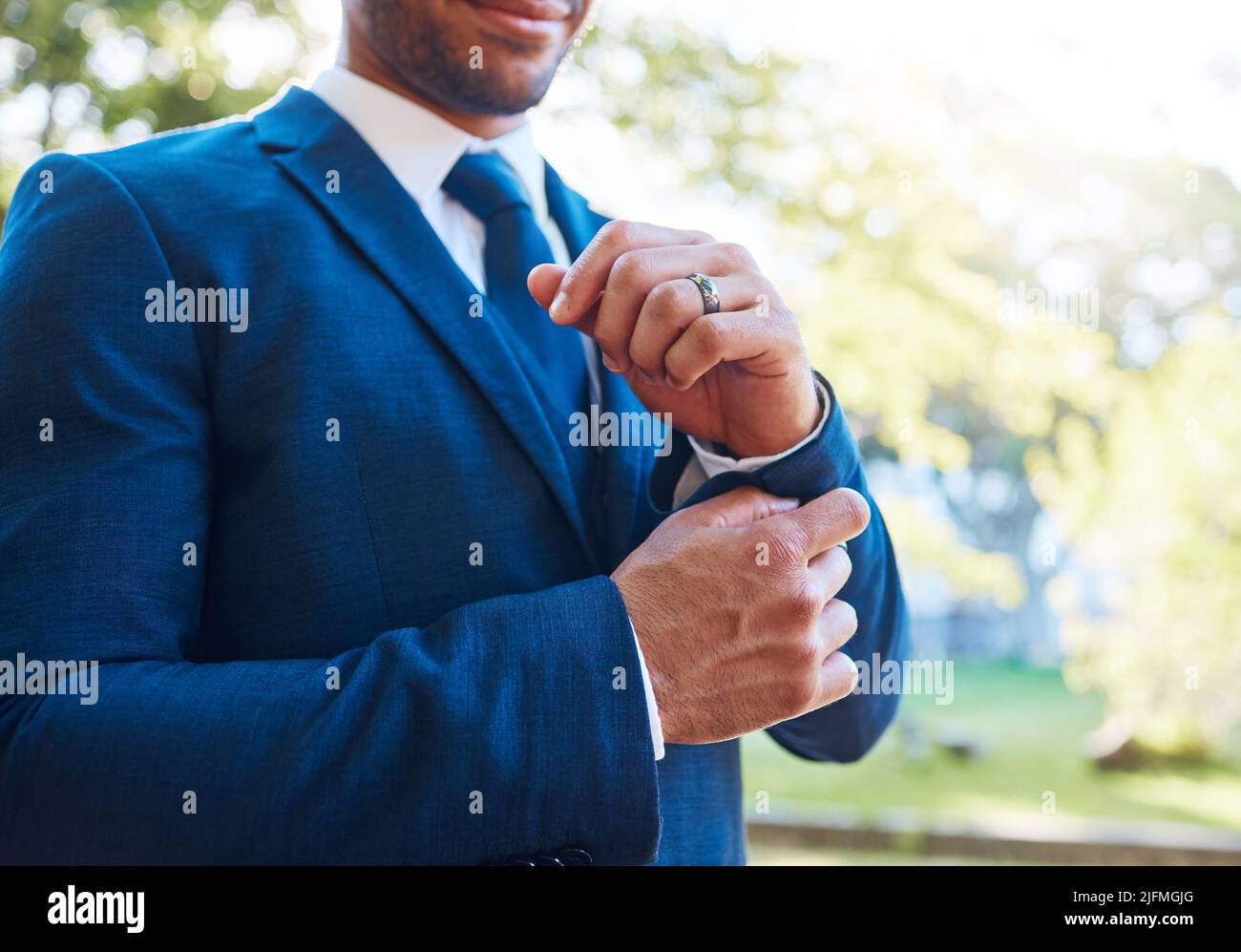 Primo piano di uno sposo che indossa un abito blu con una camicia bianca e cravatta. Sposo che regola i suoi gemelli e indossa una fascia da matrimonio mentre è in piedi Foto Stock